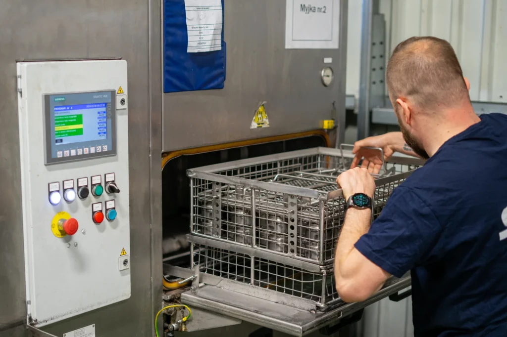 Pracownik umieszcza metalowe elementy w koszu myjki przemysłowej do wykończenia powierzchni po obróbce CNC.